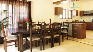 A dining area with a wooden table and chairs, a vase with flowers, and a kitchen in the background. - Lotus By Jade (Devanahalli)