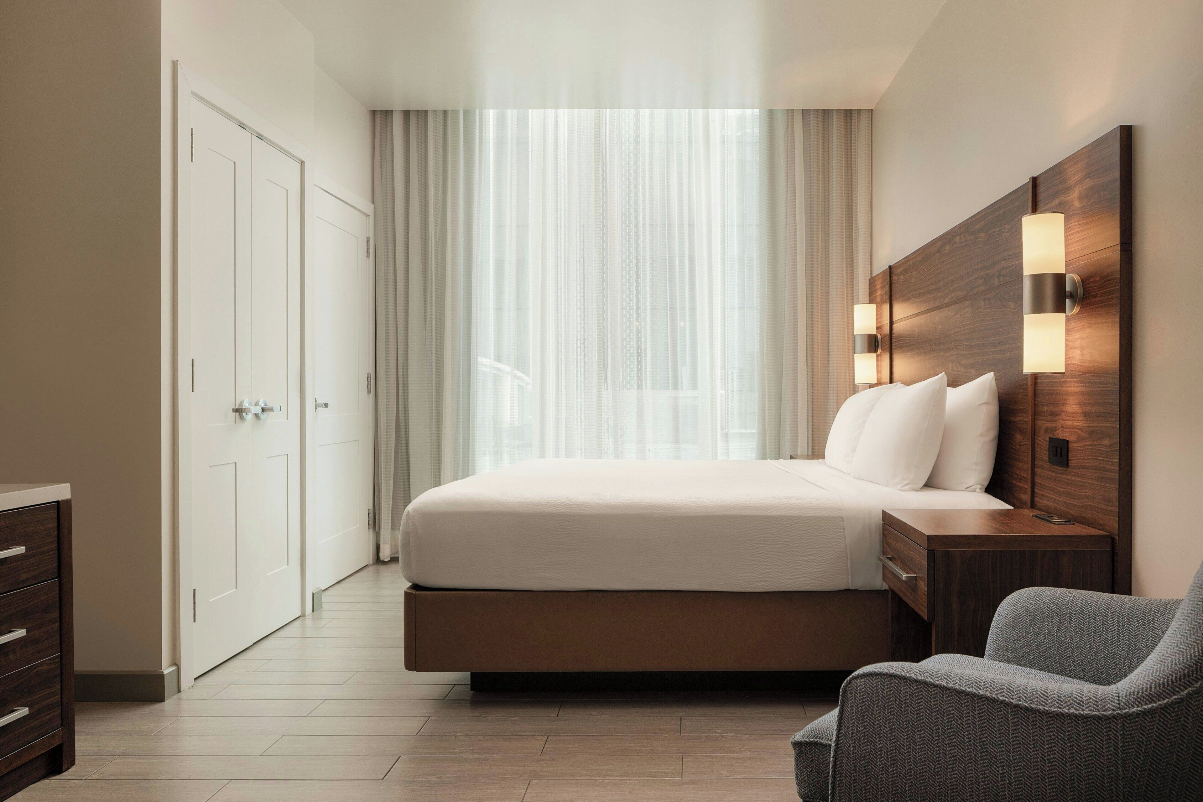 Une chambre d’hôtel avec un lit, une table de chevet, une chaise, une armoire et une fenêtre avec des rideaux.