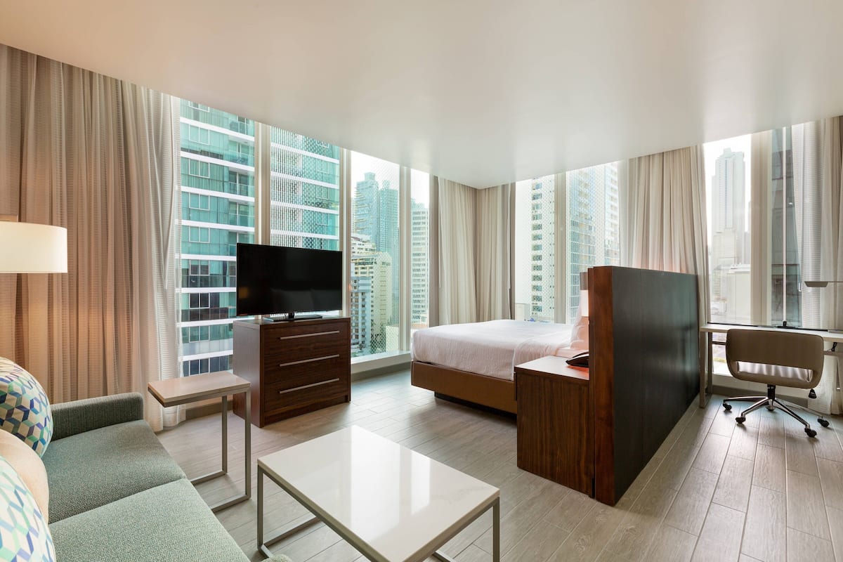 Ein Hotelzimmer mit einem Bett, einem Schreibtisch, einem Fernseher, einer Sitzecke und großen Fenstern mit Blick auf die Stadt.