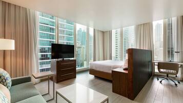 A hotel room with a bed, a desk, a TV, a sofa, and large windows with a city view.
