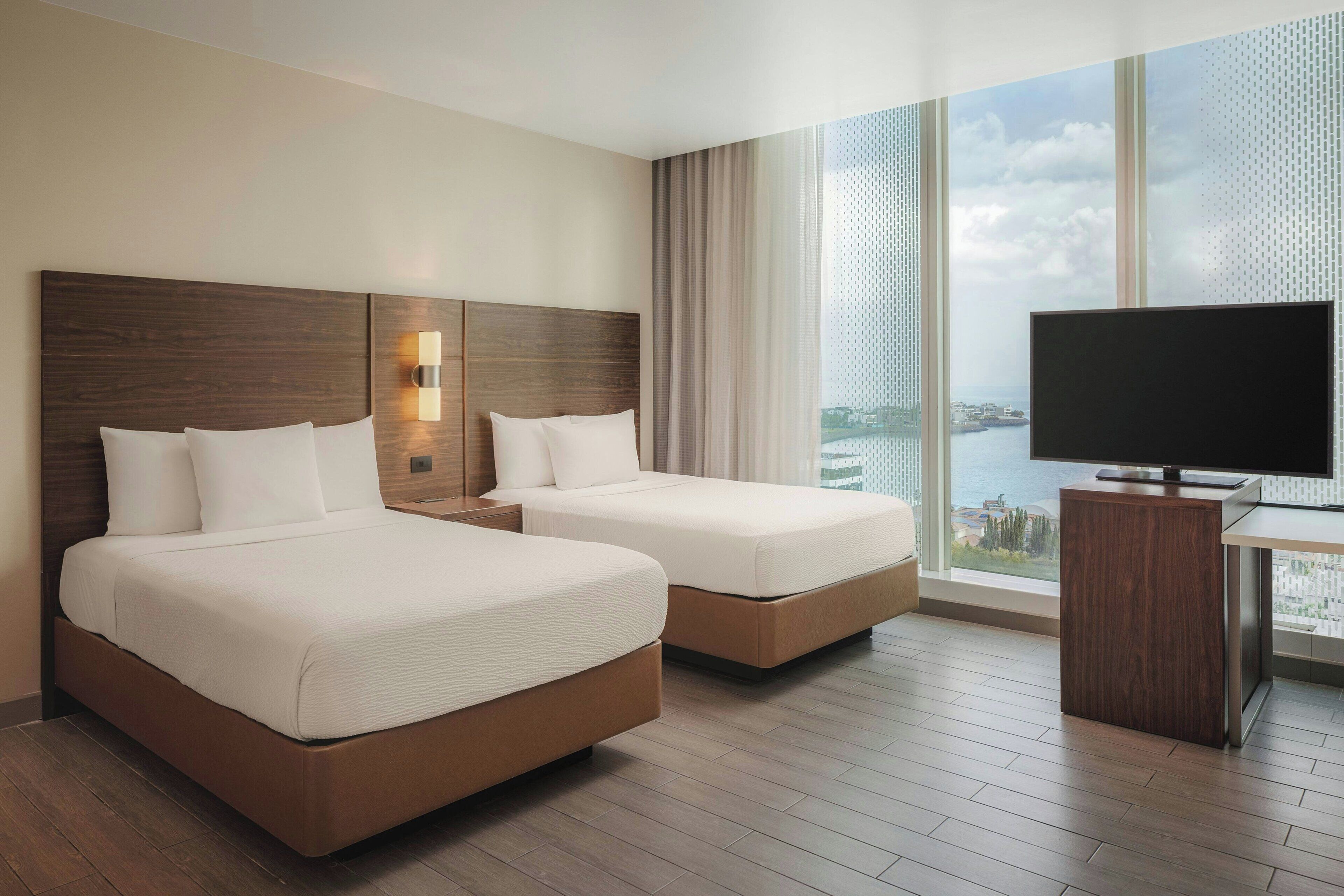 Ein Hotelzimmer mit zwei Betten, einem Fernseher, einem großen Fenster mit Stadtblick und Holzboden.