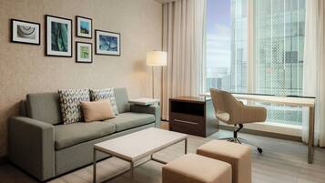 A modern hotel room with a sofa, a desk, a chair, a lamp, and framed pictures on the wall.