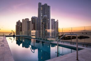 Piscina moderna de hotel con una barandilla de vidrio, rodeada de edificios altos y una vista al atardecer.