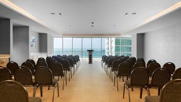 Une salle de conférence avec des rangées de chaises disposées face à un pupitre, de grandes fenêtres offrant une vue sur la mer et un sol recouvert de moquette.