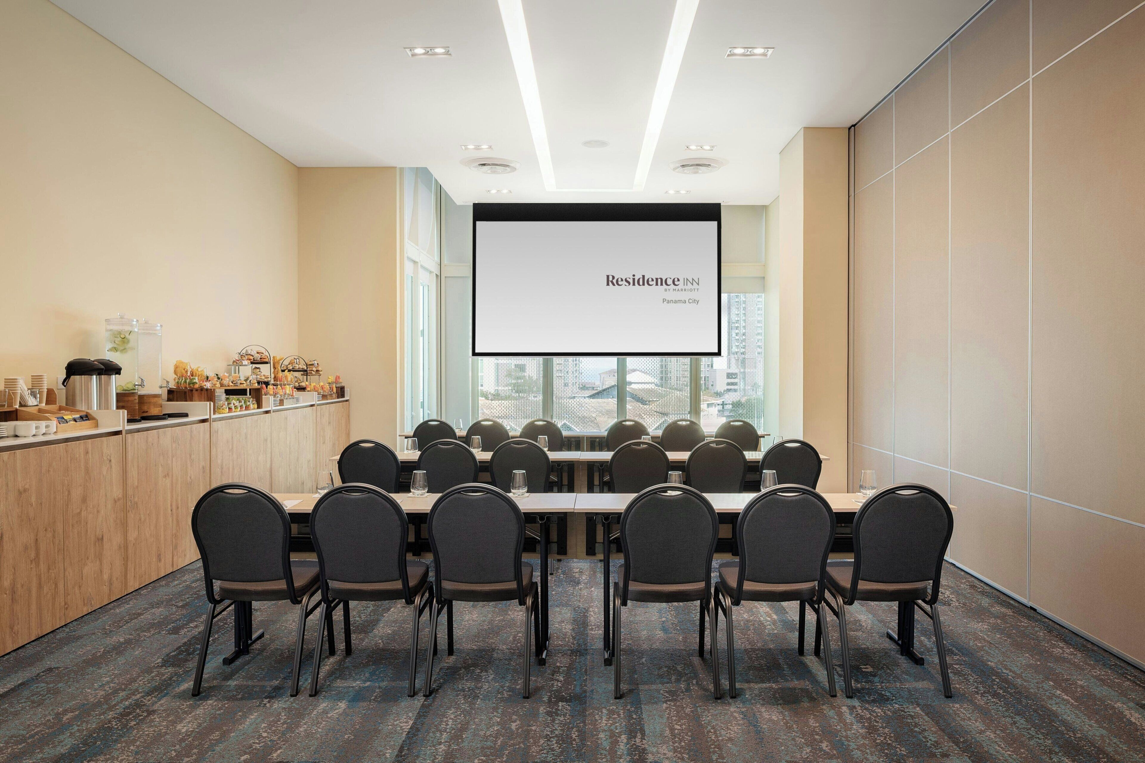 A conference room with a large screen displaying 'Residence Inn by Marriott Panama City,' a long table with chairs, a wooden counter with amenities, and large windows.