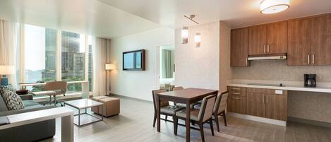 A modern hotel room with a dining area, a sofa, a TV, and a city view.