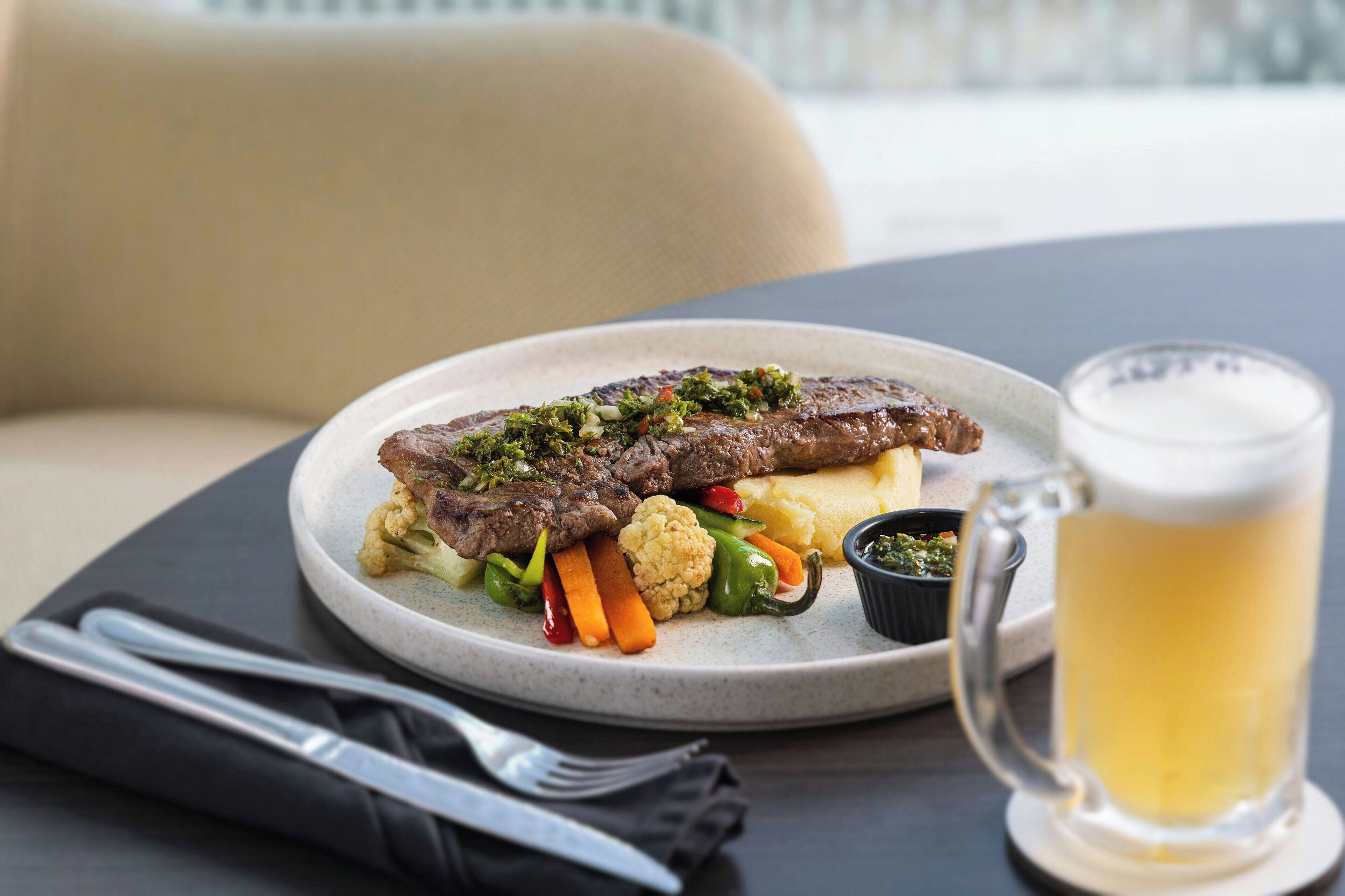 Un plato de comida con un filete, verduras y puré de papas, acompañado de una jarra de cerveza y cubiertos sobre una mesa.