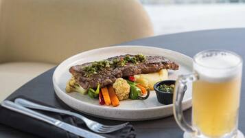 Ein Teller mit Essen, bestehend aus einem Steak, Gemüse und Kartoffelpüree, dazu ein Bierkrug und Besteck auf einem Tisch.