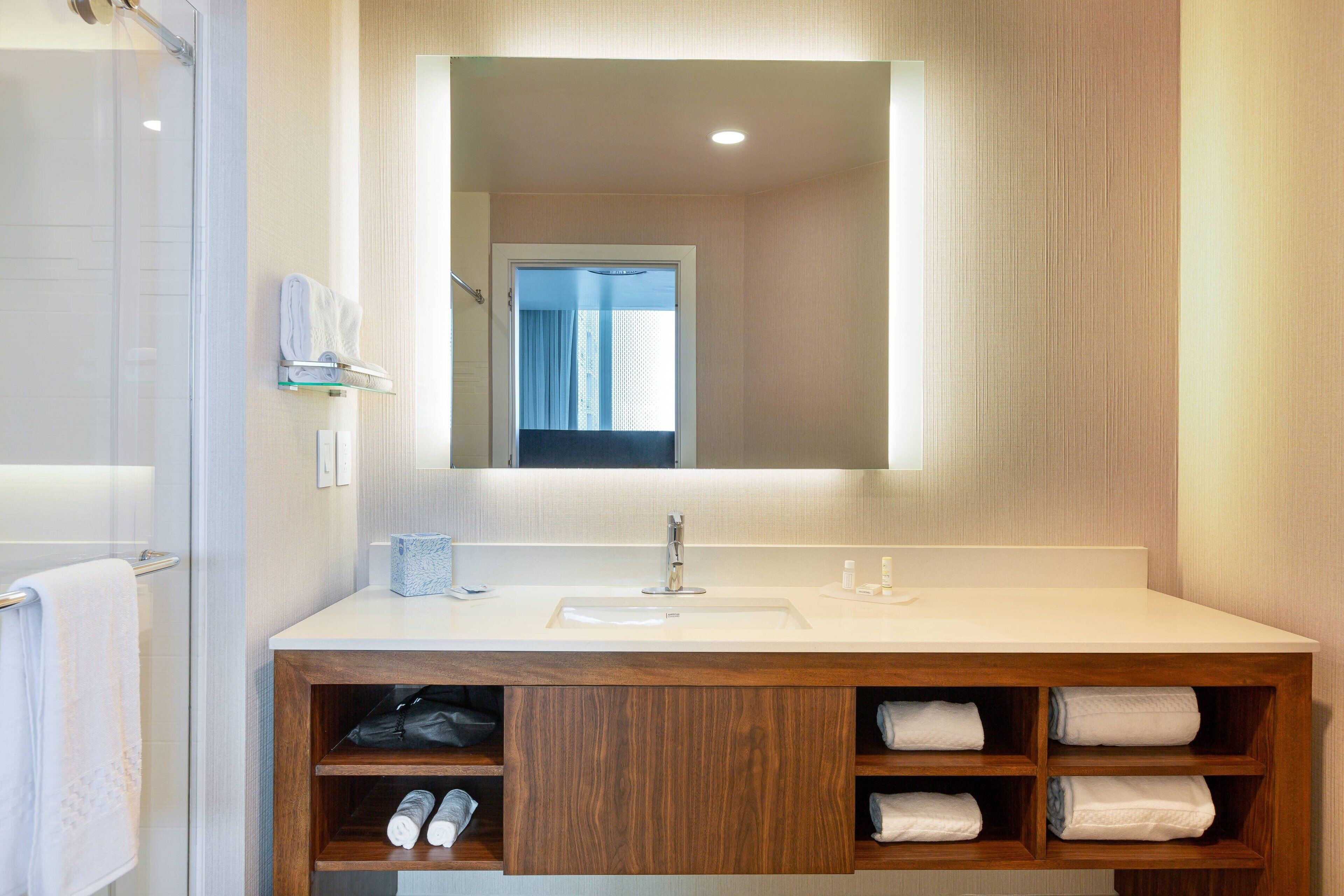 Une salle de bain moderne avec un grand miroir, un lavabo blanc et un meuble-lavabo en bois doté d’étagères pour les serviettes.