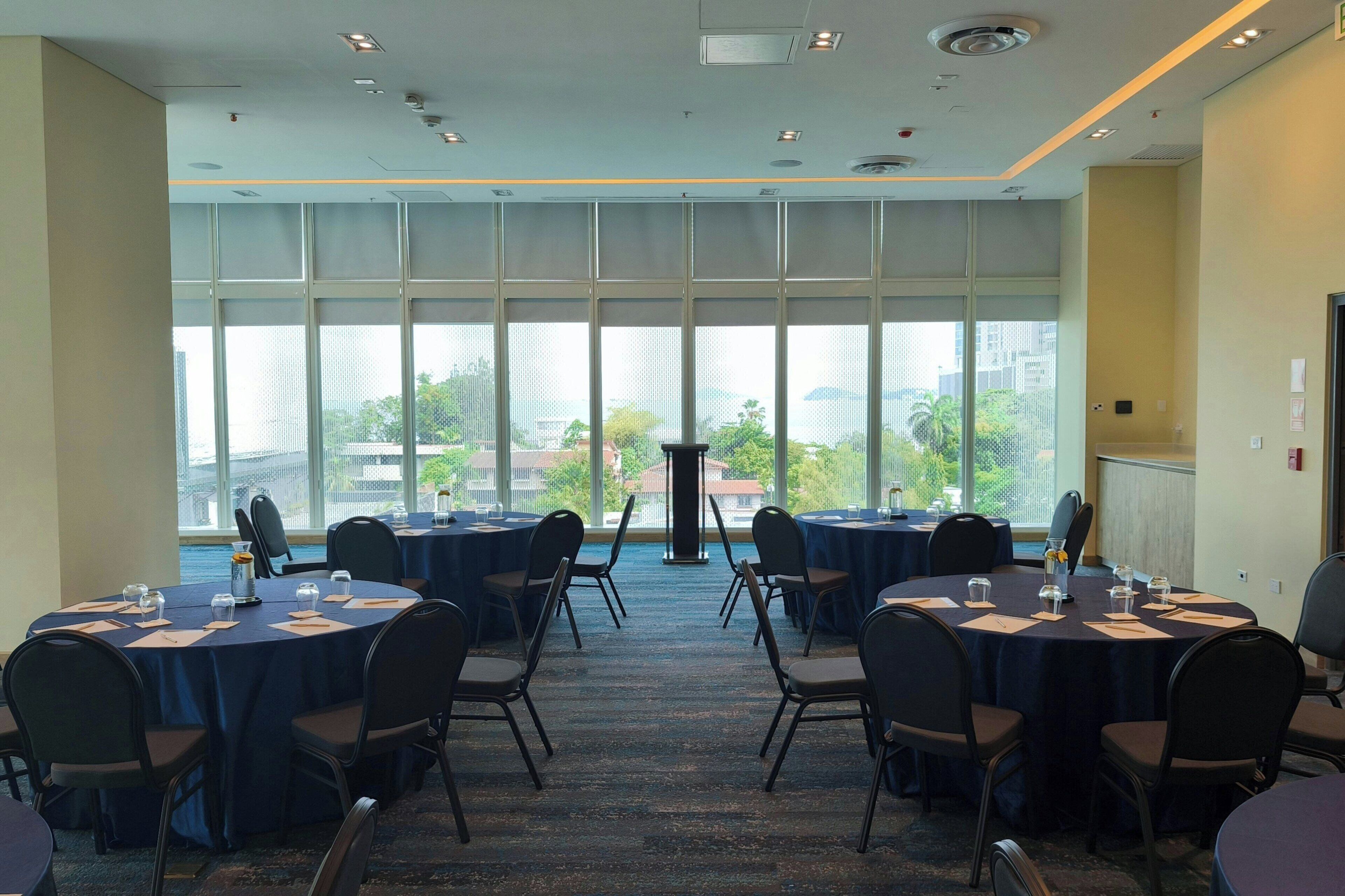 Un salón de conferencias con mesas y sillas redondas, amplias ventanas con vistas a edificios y árboles, y suelo alfombrado.