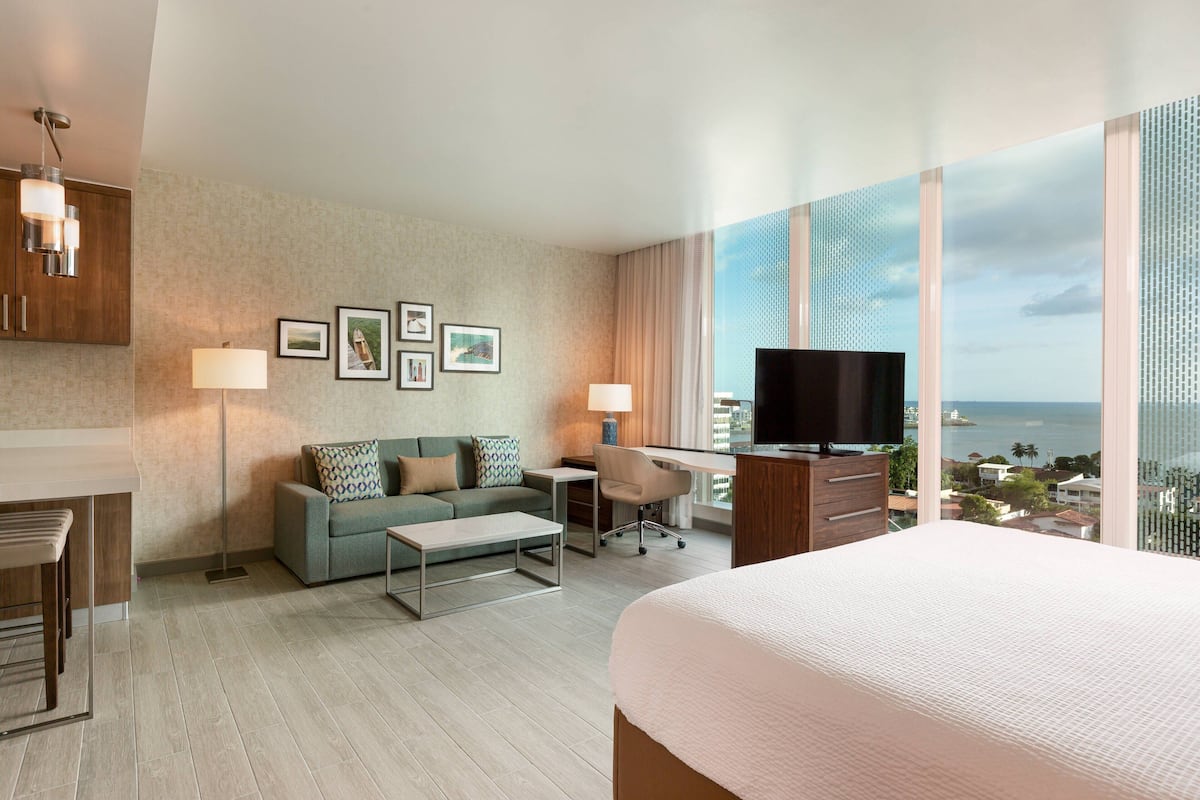 A hotel room with a bed, a desk, a sofa, and a TV. There are framed pictures on the wall and a view of the city through large windows.