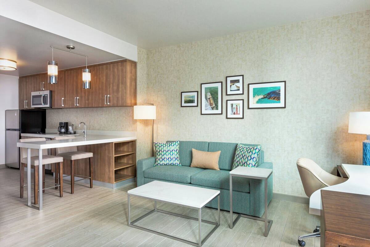 A modern kitchen with a microwave, coffee maker, and bar stools. There is a sofa with patterned cushions and a small table with a lamp.