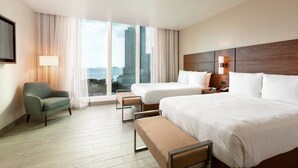 Une chambre d’hôtel avec deux lits, un canapé, un banc, une télévision et une vue sur les bâtiments.