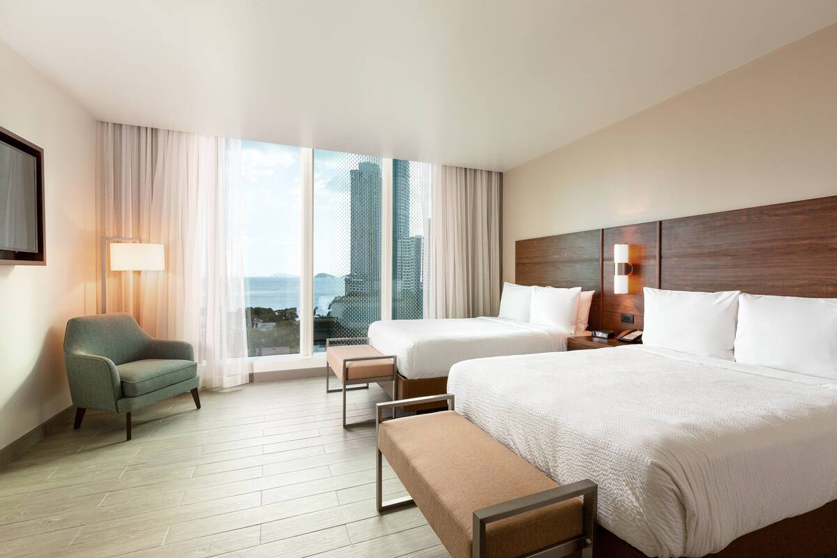 A hotel room with two beds, a sofa, a bench, a TV, and a view of buildings.