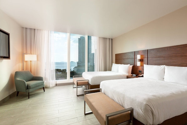 Habitación de hotel con dos camas, un sofá, un banco, una televisión y vistas a edificios.