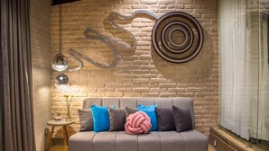 A modern living room with a grey sofa, blue and grey cushions, a decorative wall piece, and a small side table with a lamp and flowers.