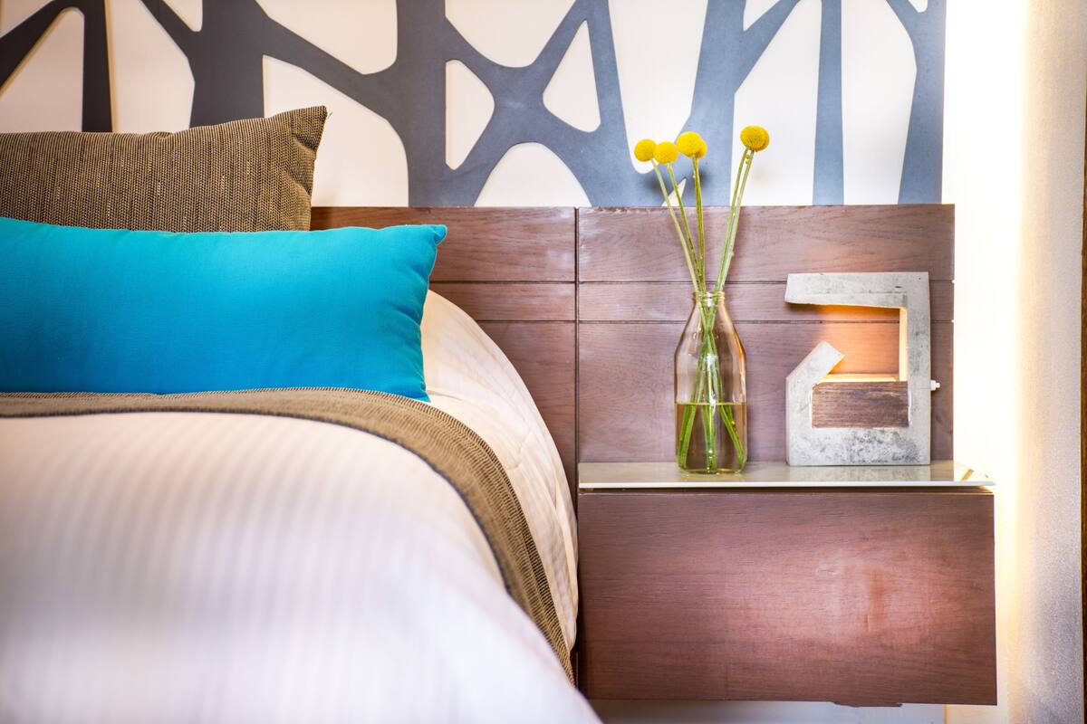 A neatly made bed with a blue pillow, a brown pillow, and a teal blanket. There is a small wooden nightstand with a vase of yellow flowers and a small lamp.