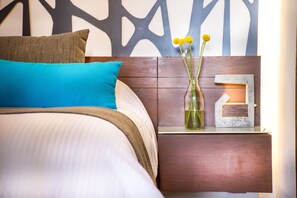 A neatly made bed with a blue pillow, a brown pillow, and a teal blanket. There is a small wooden nightstand with a vase of yellow flowers and a small lamp.