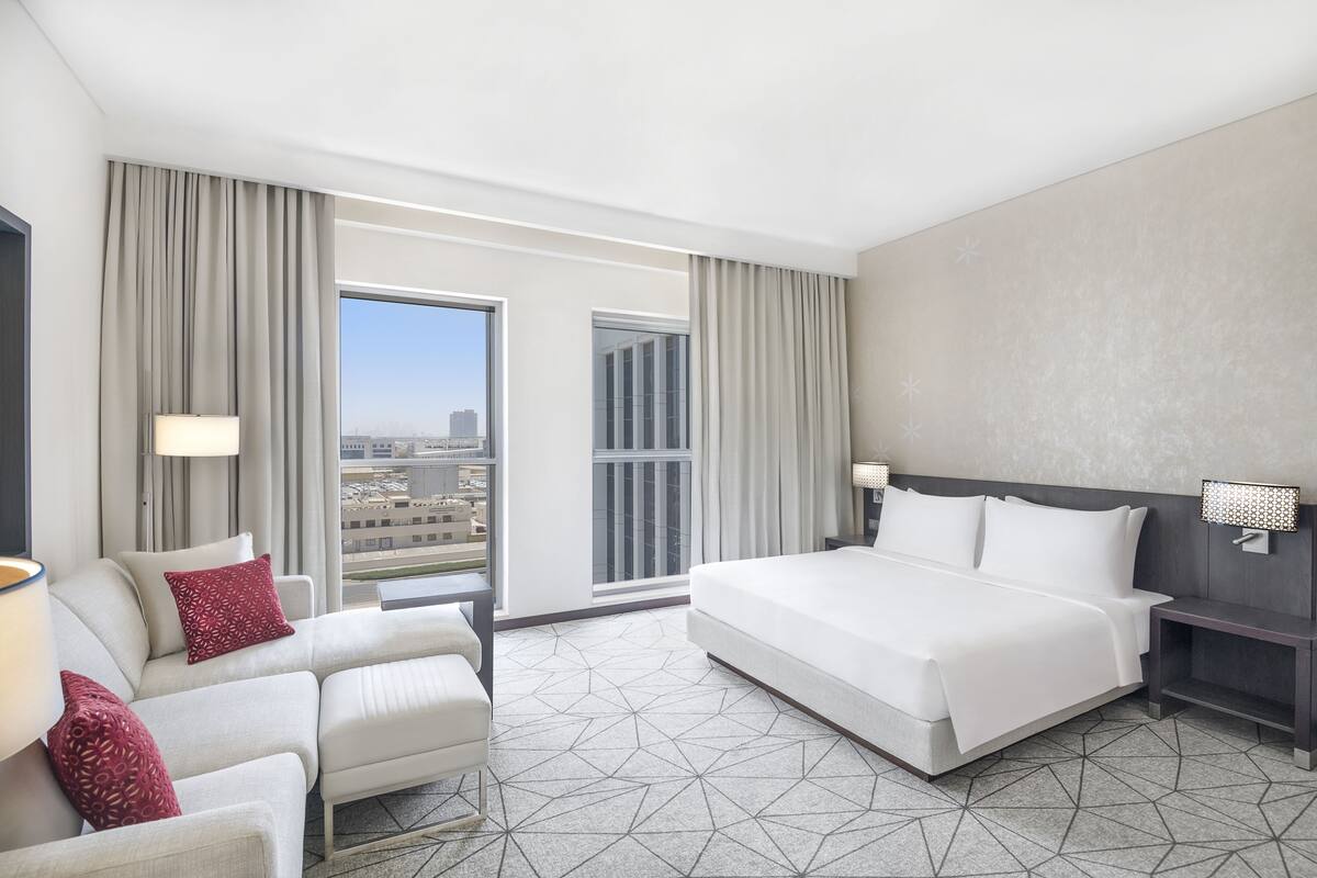 A hotel room with a large bed, a sofa, a nightstand, and a view of the city through the window.