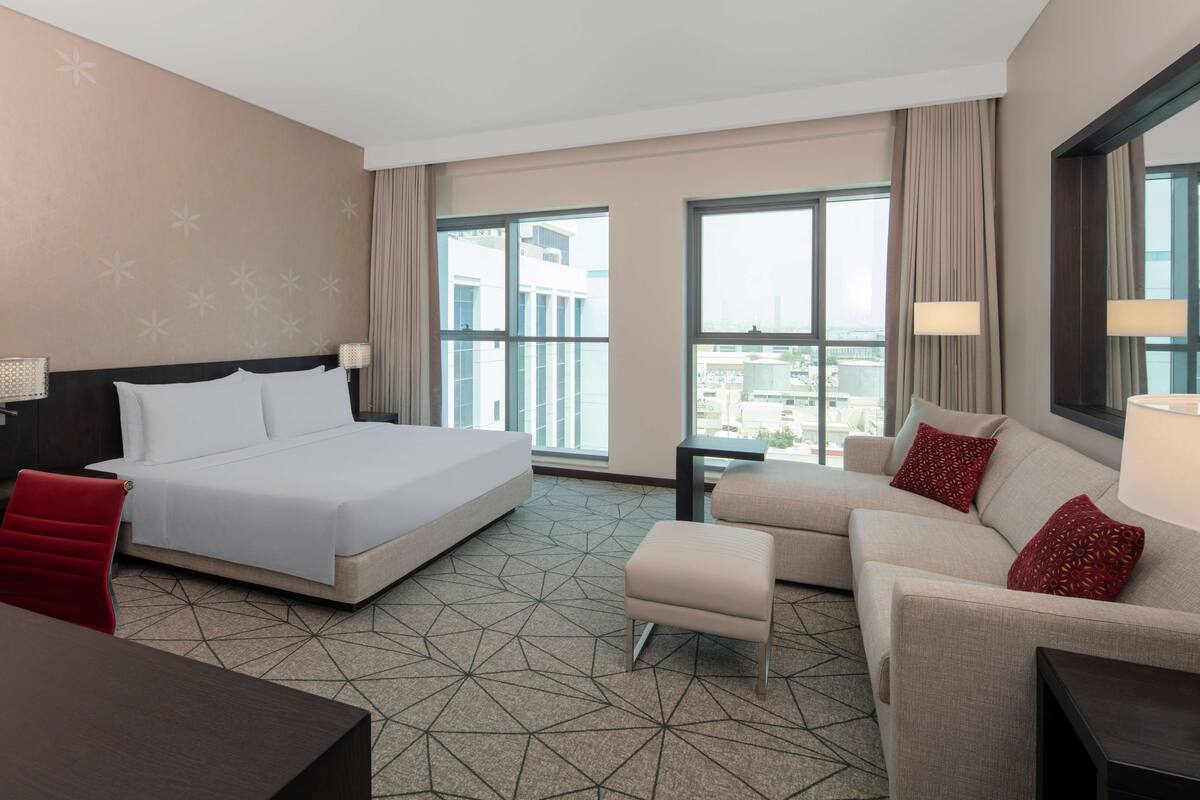 A hotel room with a large bed, a sofa, a small table, and a chair. There are large windows with a city view.
