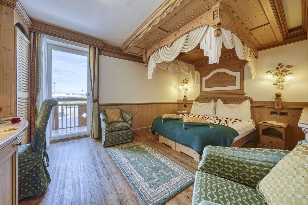 A bedroom with a wooden ceiling, a large bed, a green patterned chair, and a view through a glass door.