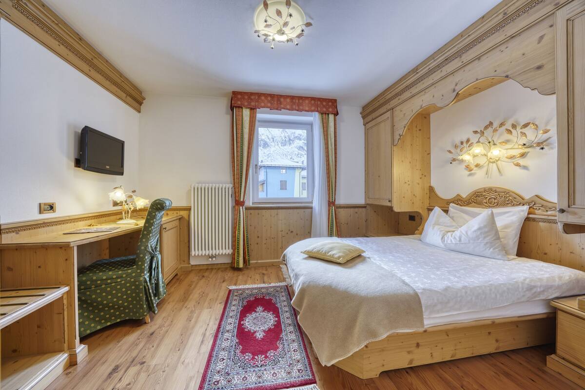 A bedroom with a wooden bed, a desk, a chair, a television, and a window with curtains.