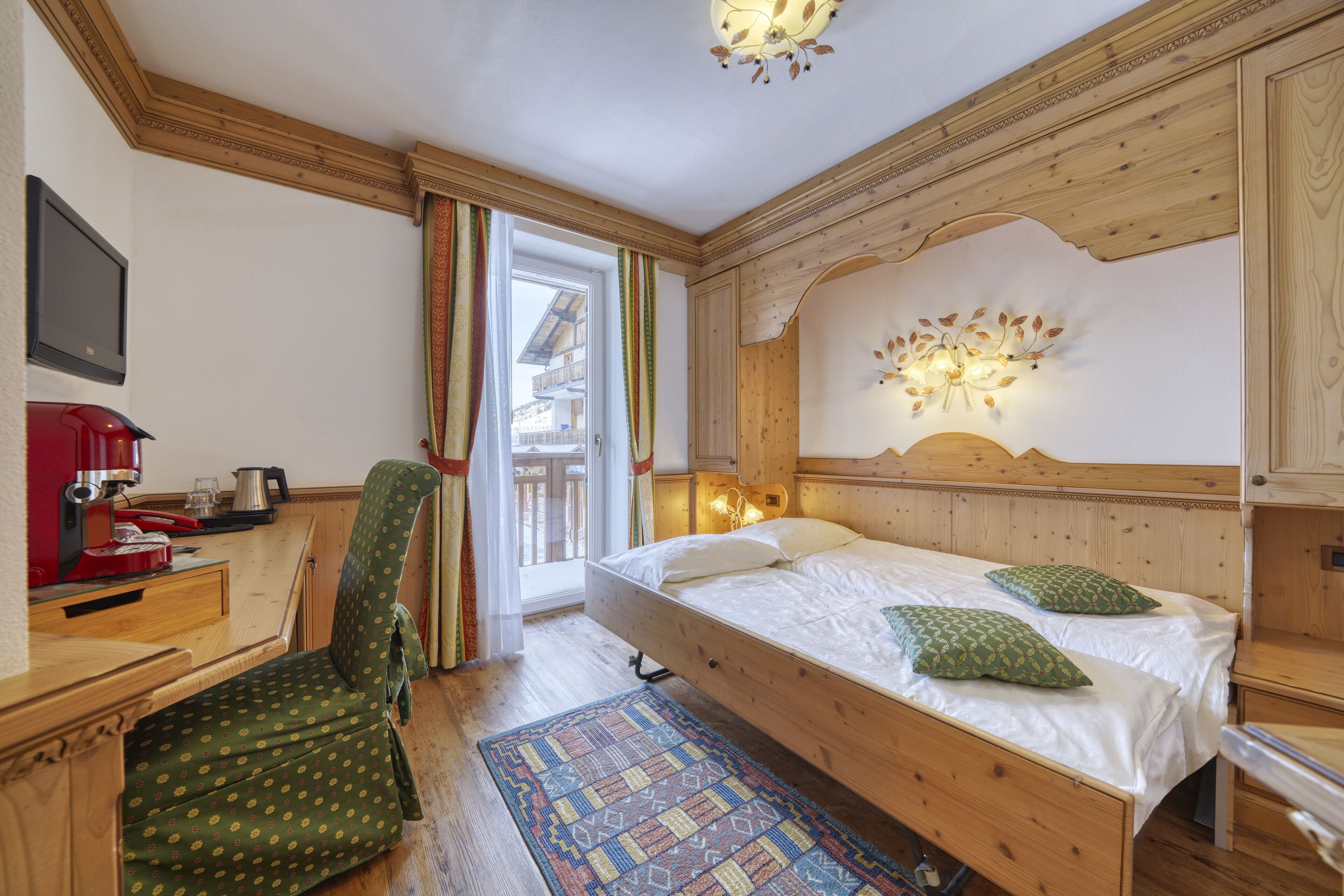 A wooden bedroom with a bed, a chair, a desk with a red coffee maker, and a window with curtains.