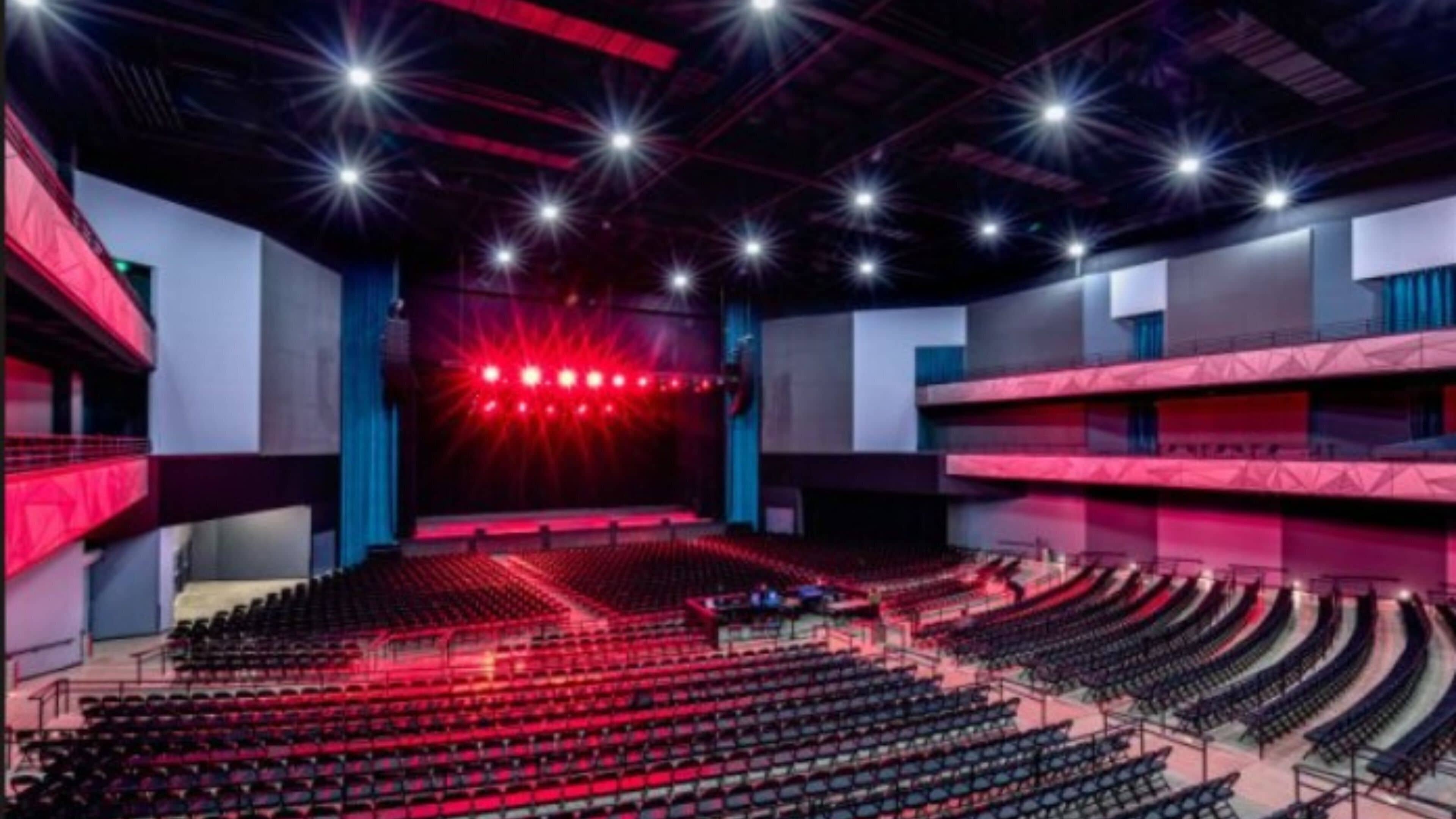Un gran auditorio interior con asientos escalonados, un escenario con iluminación roja y varias luces en el techo.