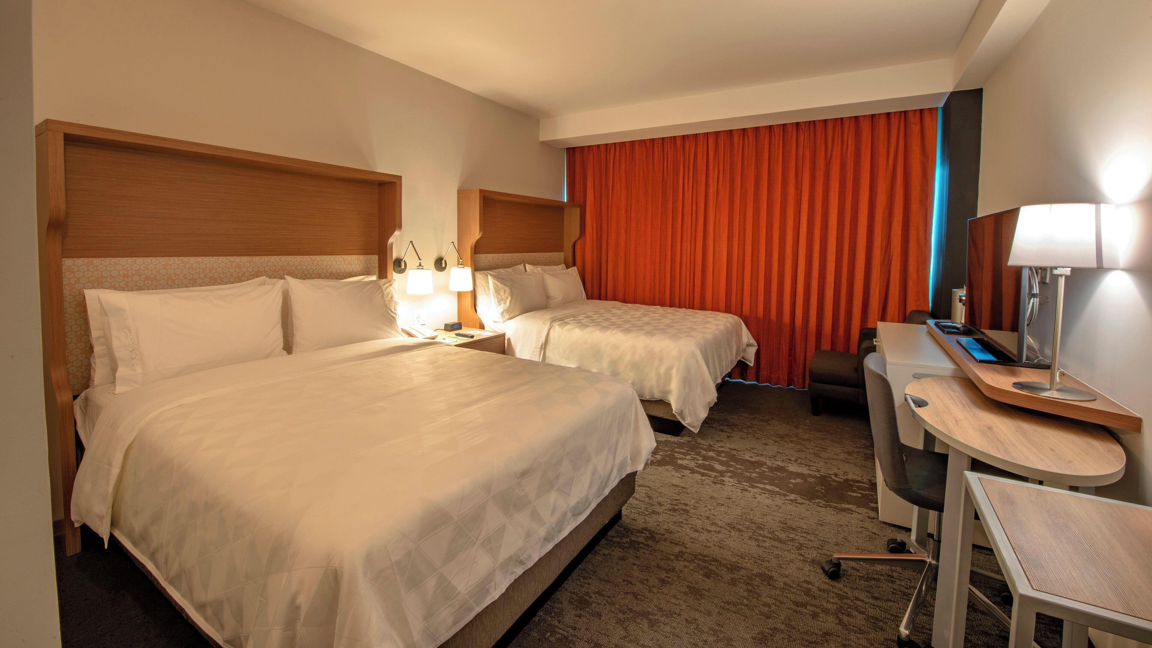 Une chambre d’hôtel avec deux lits, un bureau avec une chaise, une lampe de bureau et une grande fenêtre avec des rideaux.