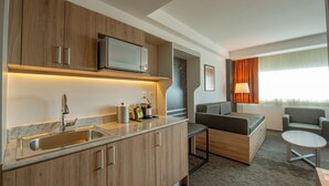 A compact hotel room with a kitchenette, a sofa, a small table, and a window with curtains.
