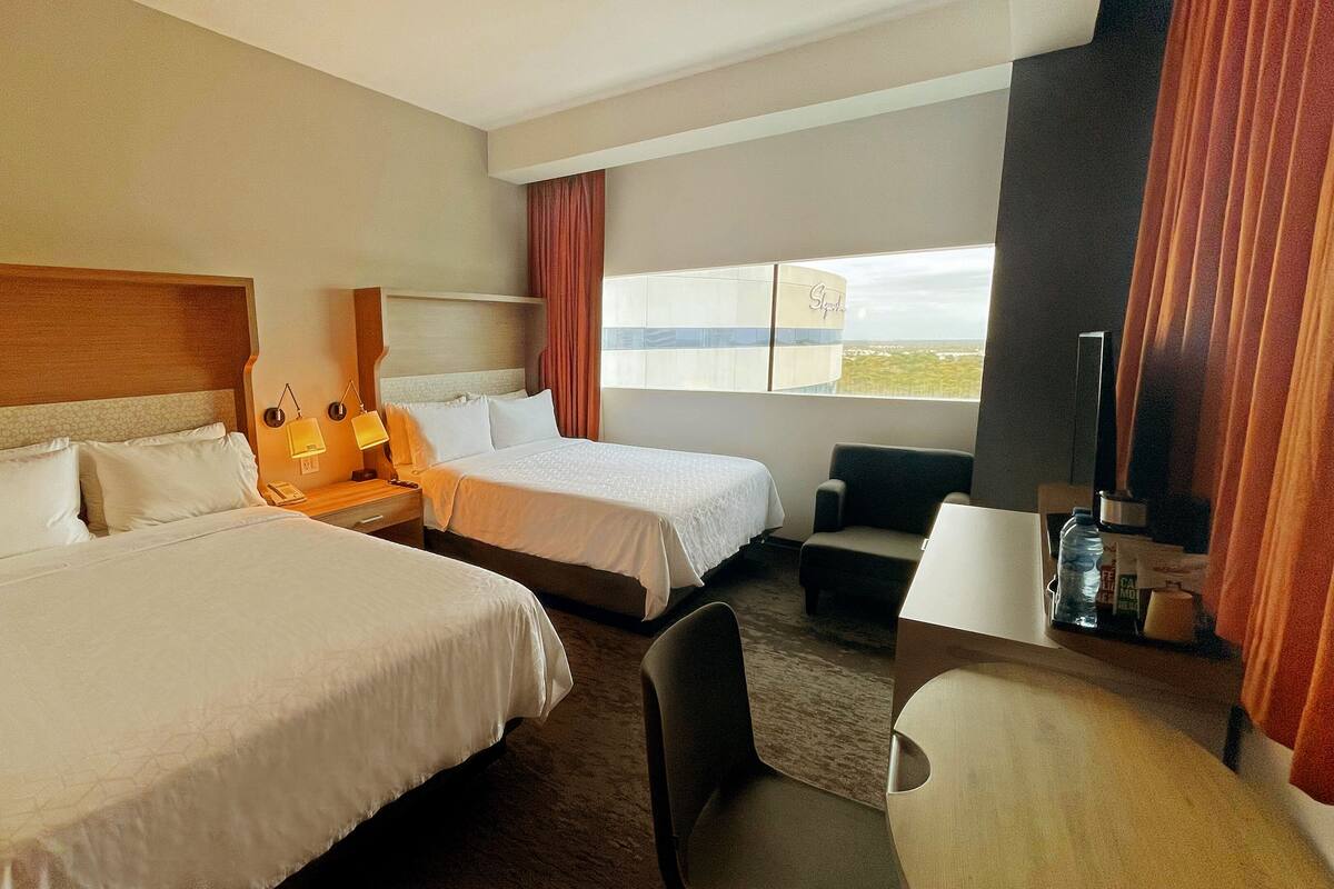 A hotel room with two beds, a desk, a chair, a TV, and a window with curtains.