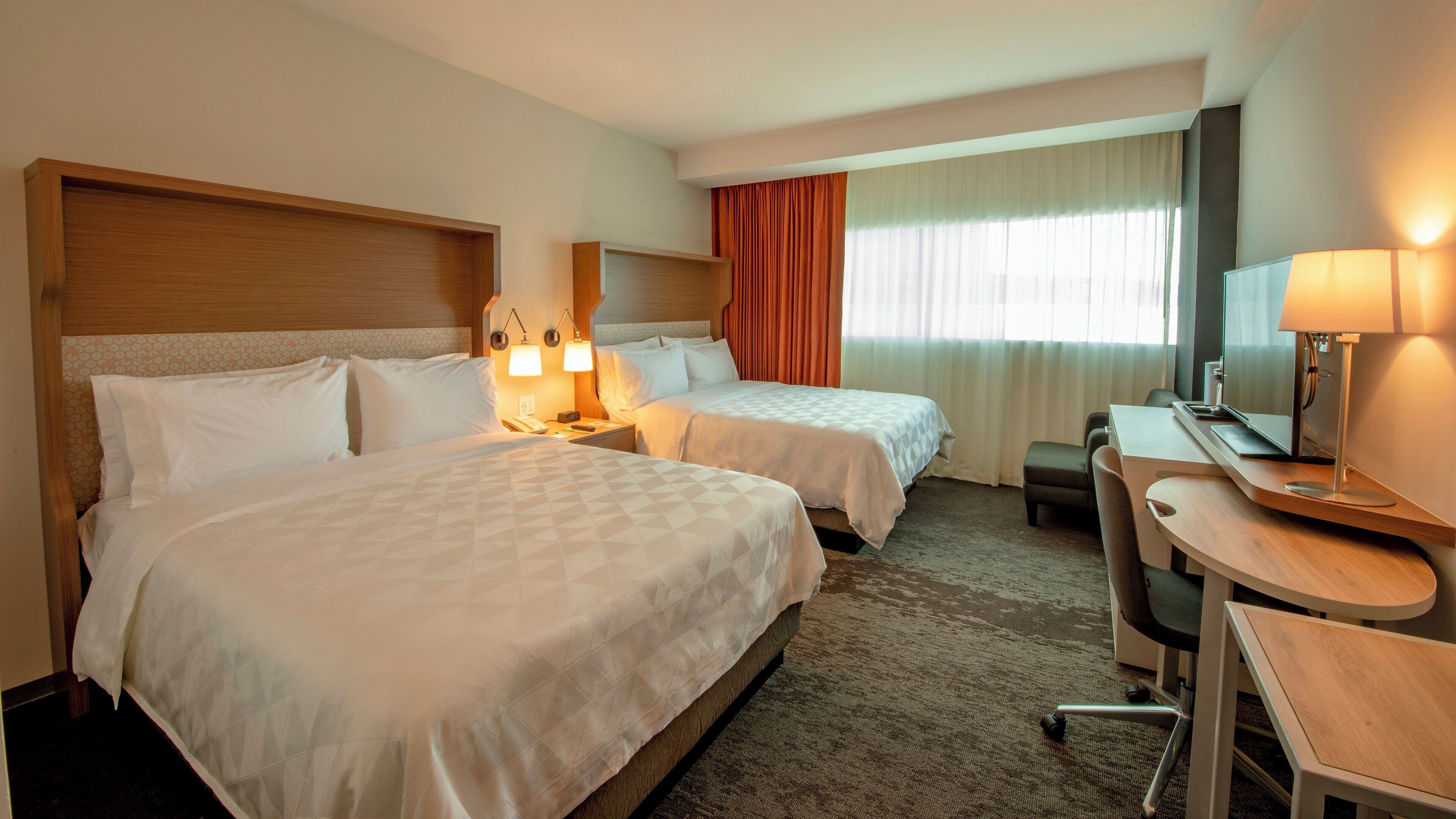 Een hotelkamer met twee bedden, een bureau, een stoel, een lamp en een groot raam met gordijnen.