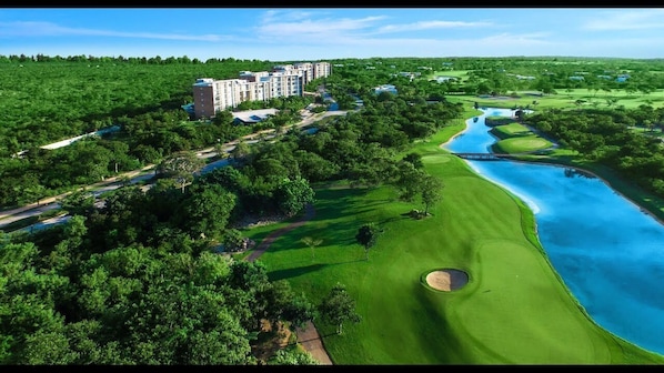 Une vue aérienne d’un terrain de golf avec un plan d’eau, entouré d’une végétation luxuriante et d’un immeuble résidentiel.