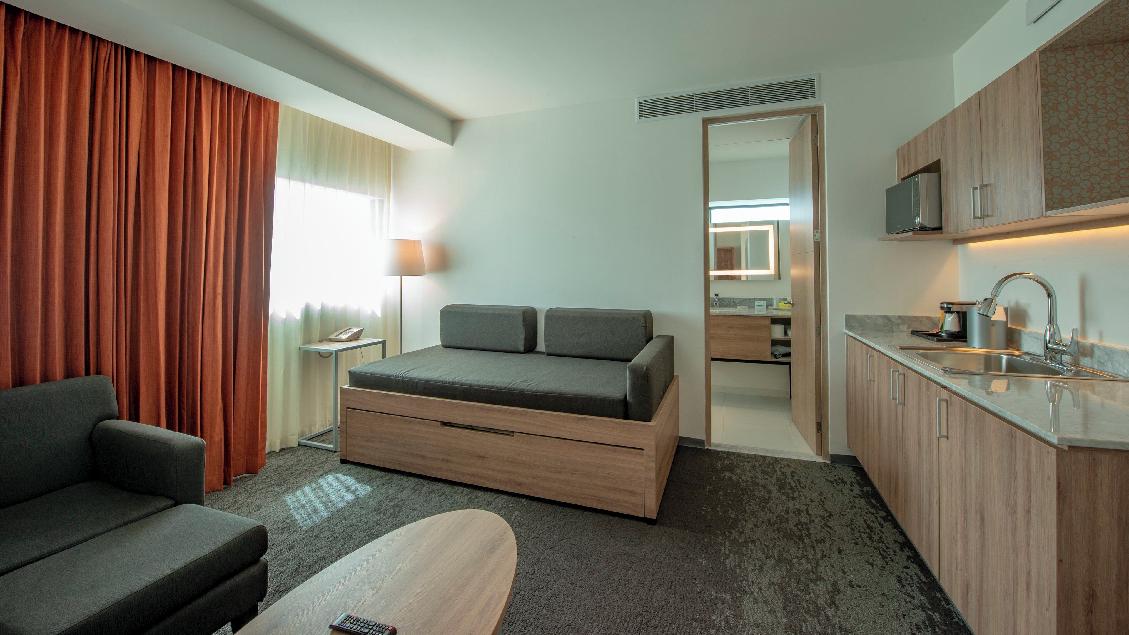 Habitación de hotel pequeña con cama, sofá, una mesita, kitchenette y baño visible a través de una puerta abierta.