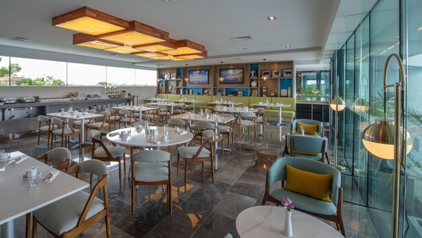 A spacious restaurant with multiple tables and chairs, a bar area, and large windows offering views of the outdoors. - Holiday Inn Hotel & Suites Merida La Isla by IHG (Mérida)