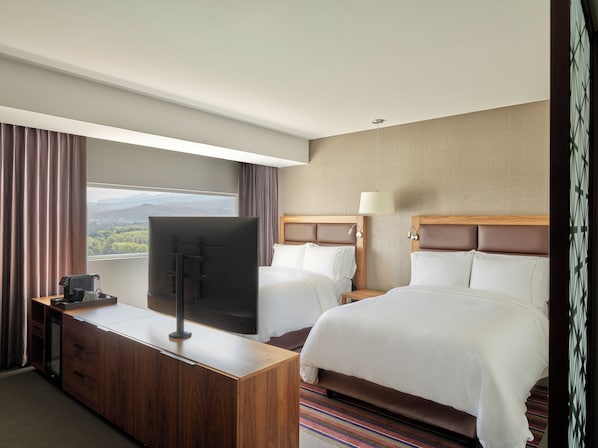 A hotel room with two beds, a TV, a desk, and a window with a view of mountains.