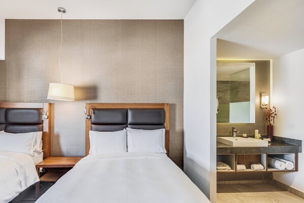 A hotel room with a large bed, a bedside table, a lamp, a mirror, and a bathroom with a sink and towel rack.