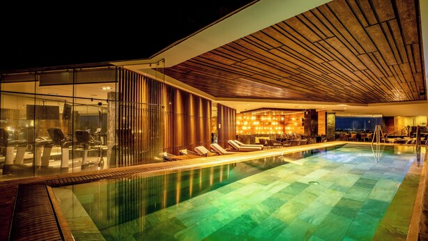 Uma área de piscina interna moderna, com paredes de vidro, espreguiçadeiras e um espaço bar.