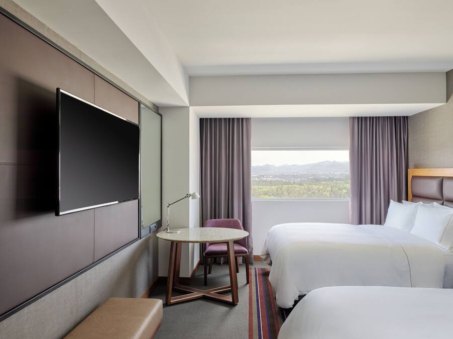 A hotel room with two beds, a TV, a desk, and a view of a cityscape.