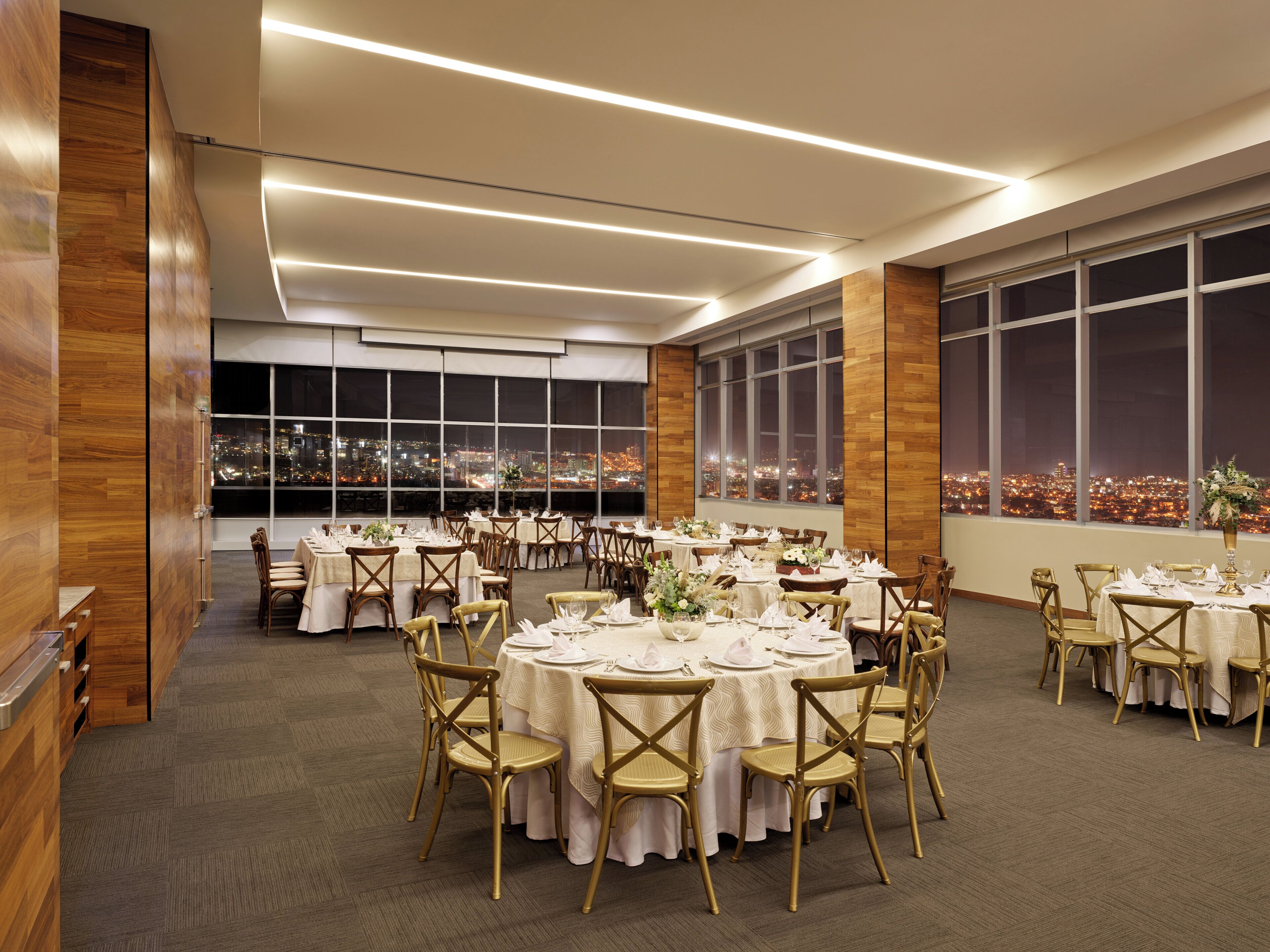 A spacious banquet hall with multiple round tables set for dining, large windows offering a city view, and a modern ceiling design.