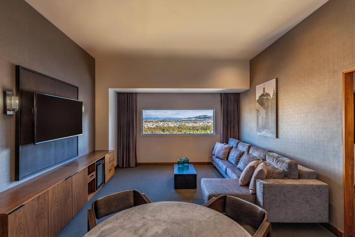 Une chambre d’hôtel moderne dotée d’une télévision à écran plat, d’un canapé, d’une table à manger et offrant une vue sur le paysage urbain grâce à une grande fenêtre.