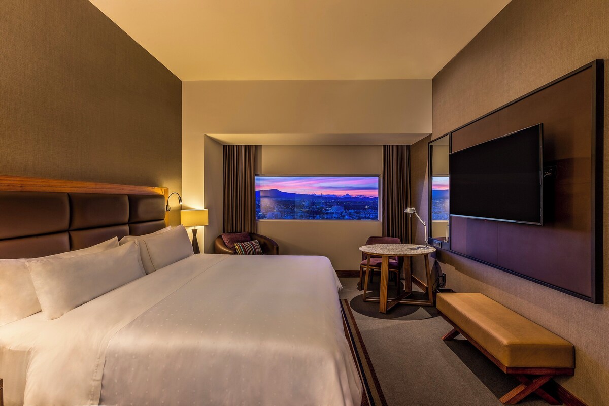Habitación de hotel con una cama grande, un televisor en la pared, una pequeña mesa redonda y un banco.