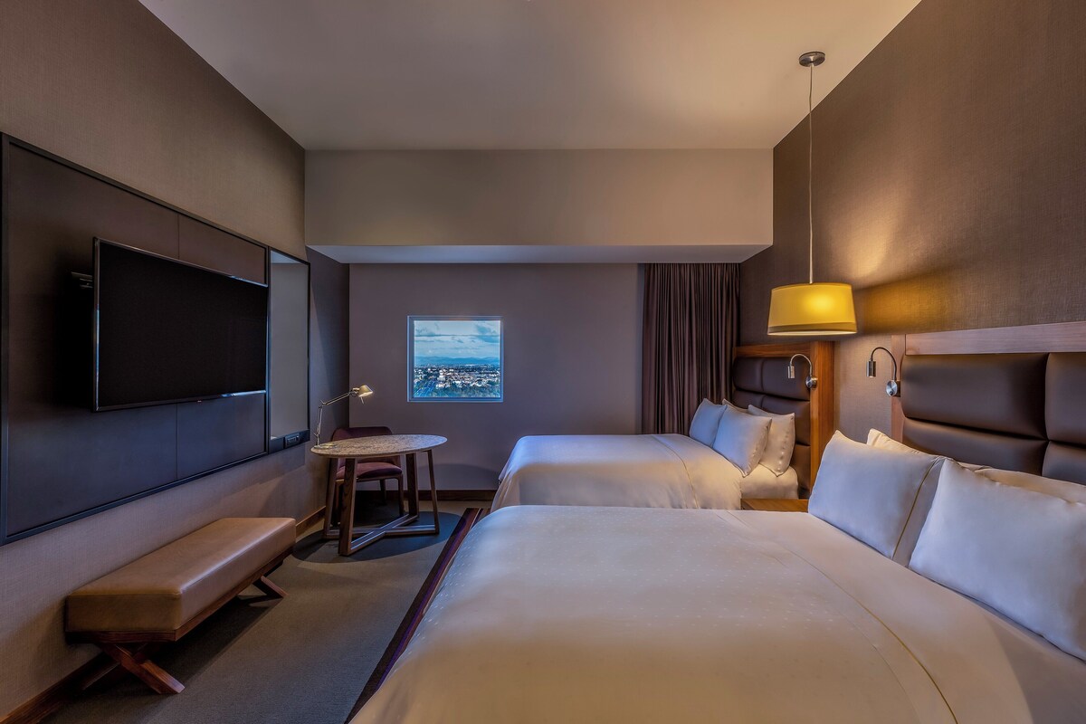 Habitación de hotel con dos camas, televisión, escritorio y ventana con vista a la ciudad.