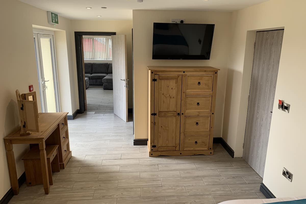 Habitación de hotel con un escritorio de madera, un armario, un televisor montado en la pared y vista al área de estar por una puerta abierta.