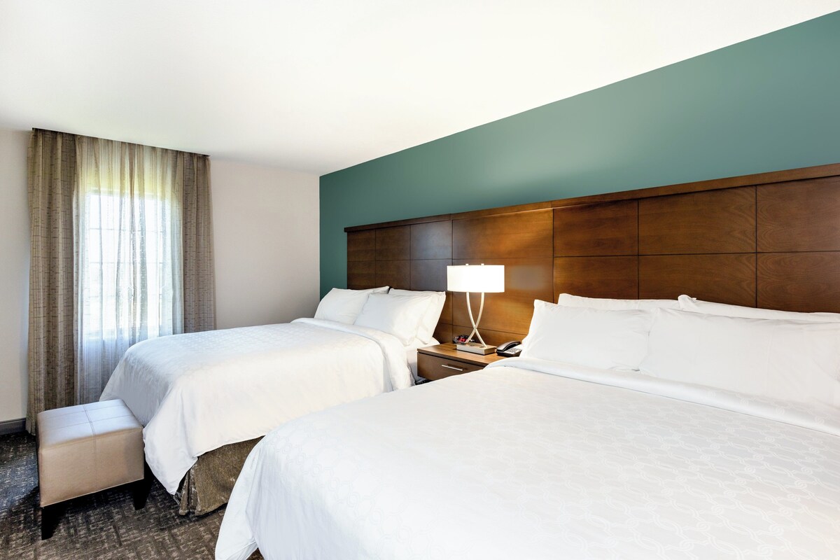 Una habitación de hotel con dos camas, una mesita de noche con lámpara, una pequeña mesa y una ventana con cortinas.