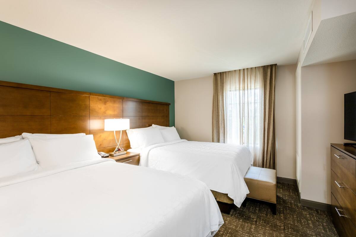 Habitación de hotel con dos camas, cabecera de madera, mesita de noche con lámpara, un pequeño banco, televisor y ventana con cortinas.
