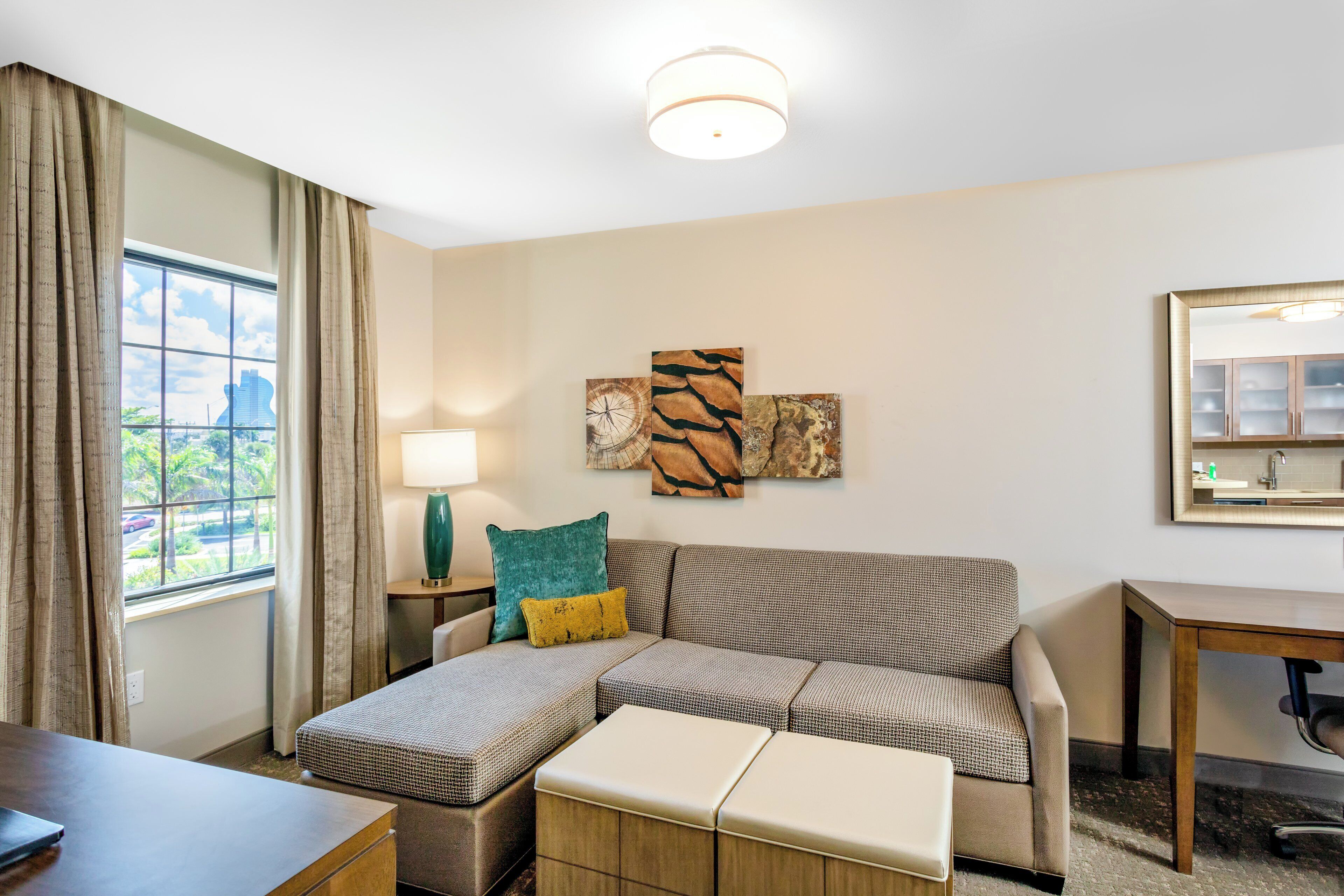 A hotel room with a sectional sofa, a small table, a lamp, and a large window with curtains.