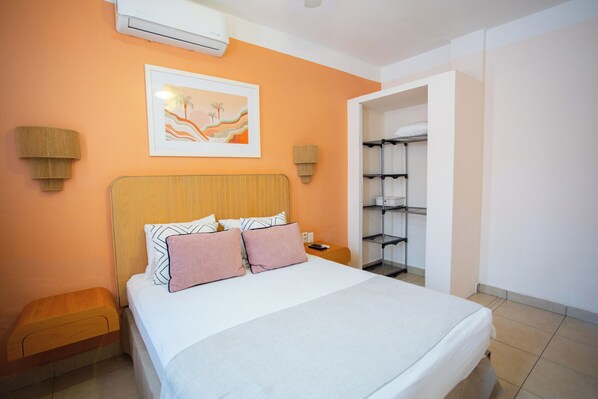 Un dormitorio ordenado con cama, mesitas de noche, aire acondicionado instalado en la pared, una obra de arte enmarco y una estantería.