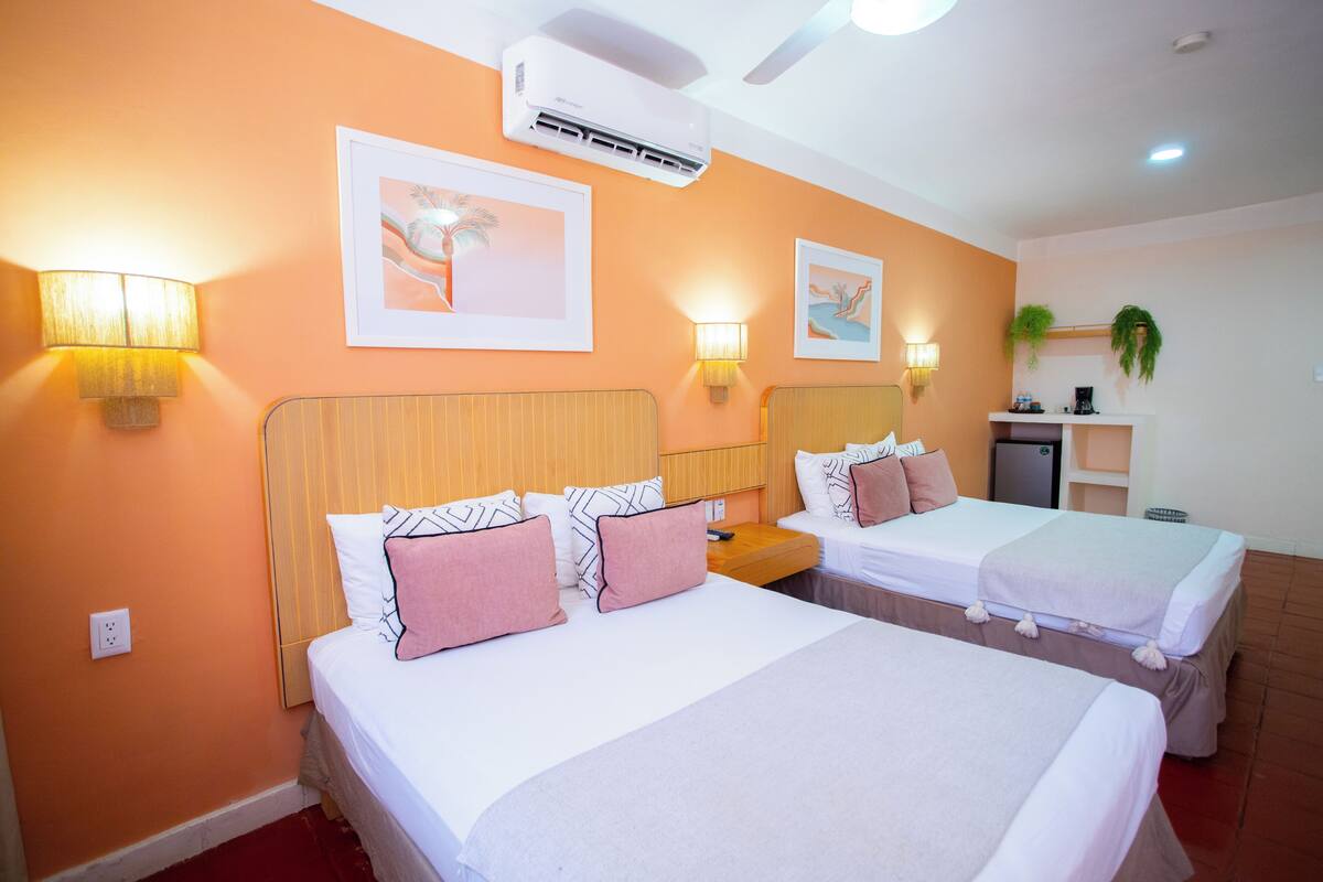 A hotel room with two beds, a ceiling fan, an air conditioner, wall-mounted lamps, and framed artwork.