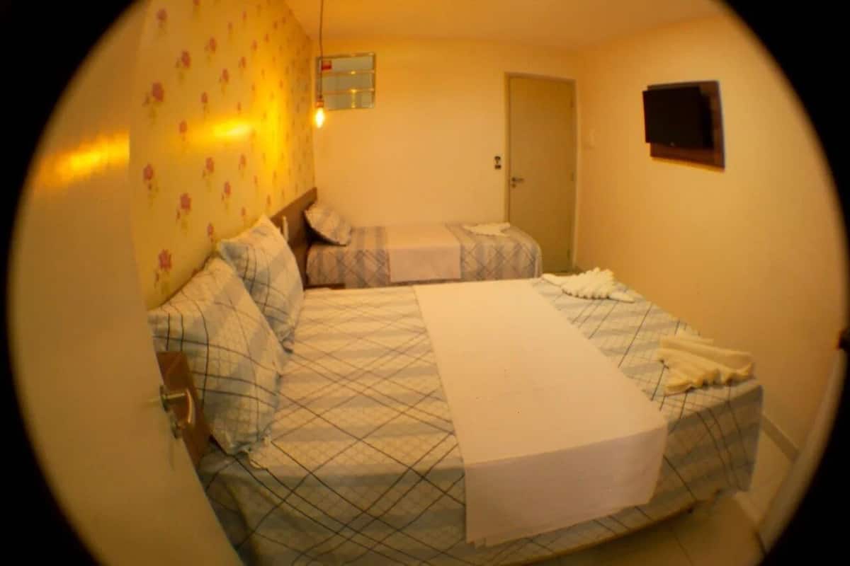 Una habitació amb dos llits, una porta, un finestral petit i una televisió penjada a la paret.